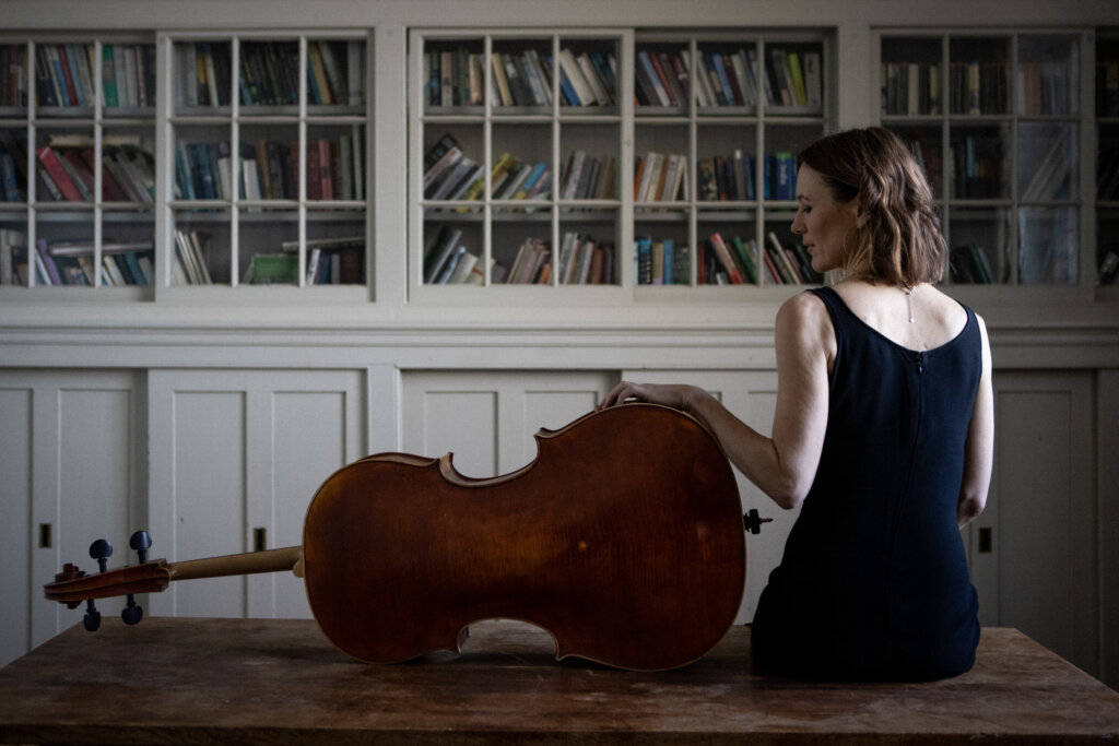 Musician sits next to cello with a bookshelf in the background
